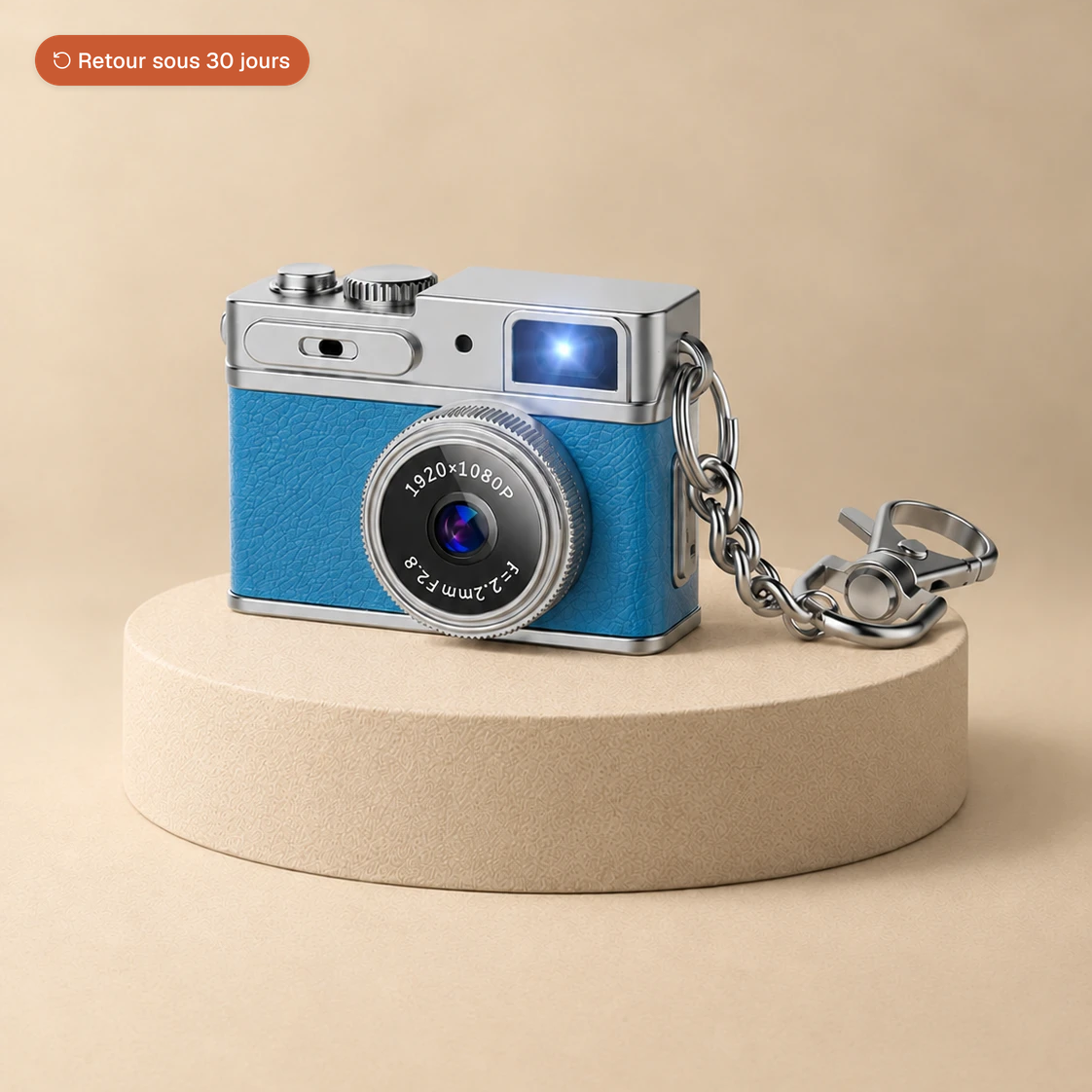 Mini appareil photo rétro bleu avec sa chaîne porte-clés, posé sur un socle beige